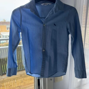 Blå skjorta från H&M med knappar - Snygg blå skjorta från H&M i storlek 134. Skjortan har klassisk krage, långa ärmar och knäppning framtill. Tillverkad i mjuk bomull och har en liten broderad detalj på bröstet. Perfekt för dig som gillar stilrena och enkla plagg.