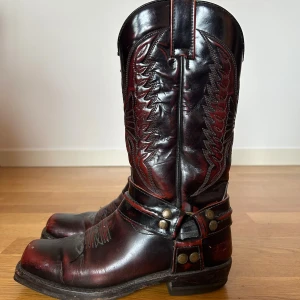 Svarta och vinröda cowboy boots Sendra - Riktigt snygga cowboy boots från Sendra i svart och vinrött skinn. Klassisk western-design med detaljerade broderier och metallnitar runt ankeln. Hög skaft och rund tå, perfekt för dig som vill sticka ut med en cool westernstil.