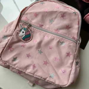 Rosa ryggsäck med stjärnor och enhörning - Söt ljusrosa ryggsäck med glittriga stjärnor i rosa och silver, samt ett gulligt enhörningsmärke på dragkedjan. Väskan har justerbara axelband och ett stort fack med extra ytterficka framtill. Perfekt för dig som gillar färgglada detaljer och magiska motiv.