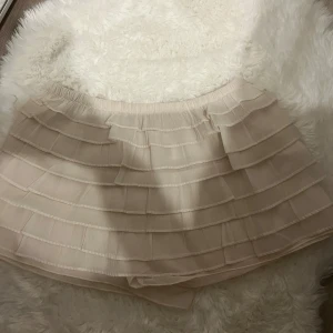 Beige volangshorts med resårmidja - Supersöta korta short i beige med flera lager volanger och elastisk midja. Kjolen har en lätt och luftig känsla, perfekt för dig som gillar en feminin och trendig stil. Passar snyggt till både sneakers och boots.