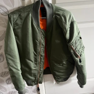 Grön bomberjacka Alpha Industries M - Säljer en klassisk grön bomberjacka från Alpha Industries i storlek M. Jackan har orange foder, ribbade muddar, dragkedja i kopparfärg och ficka med dragkedja på ärmen. Materialet är slitstark polyester och jackan har en riktigt clean och tidlös look.