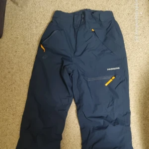 Mörkblå täckbyxor från Didriksons - Mörkblå täckbyxor från Didriksons med vattentätt material och gula detaljer vid fickorna. Byxorna har dragkedja framtill och praktiska fickor, perfekta för kalla och blöta dagar. Snygg och funktionell design för dig som gillar att vara ute.