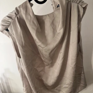 Beige blus från Gina Tricot - Säljer en beige blus från Gina Tricot i storlek 32. Blusen har en mjuk, draperad krage och är ärmlös med lätt rynkade axlar. Materialet är slätt och har en elegant känsla, perfekt för att lyfta din outfit. Enkel att matcha med både jeans och kjol.