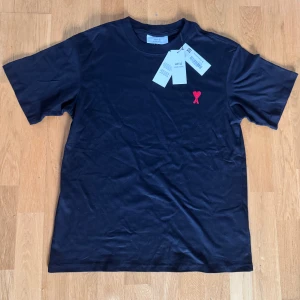 Svart t-shirt från Ami med rött hjärta - Svart t-shirt från Ami Alexandre Mattiussi i mjuk bomull. Klassisk passform med korta ärmar och rund hals. På bröstet finns ett broderat rött hjärta med A och på ryggen är Ami-loggan broderad. Perfekt för en clean och stilren look.