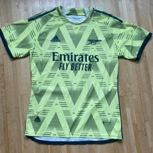 Arsenal gul matchtröja Adidas M - Säljer en Arsenal fotbollströja från Adidas i gul färg med mörkblå detaljer. Tröjan har ett grafiskt zigzag-mönster, korta ärmar och rund hals. Emirates Fly Better-logga på bröstet och klubbens kanon-emblem. Tillverkad i lätt, ventilerande material.