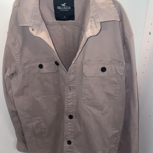 Beige overshirt från Hollister - Snygg beige overshirt från Hollister med klassisk krage och svarta knappar. Jackan har två bröstfickor med lock och knapp samt raka sidfickor. Perfekt lager-på-lager-plagg för en chill och stilren look.