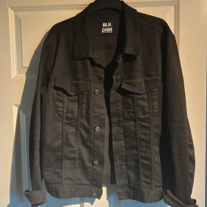 Svart jeansjacka från BLK DNM - Stilren svart jeansjacka från BLK DNM med klassisk krage, knäppning framtill och två bröstfickor med lock. Jackan har raka linjer och silverfärgade knappar. Perfekt för dig som gillar en clean och tidlös look.