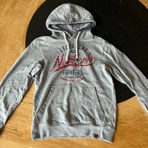 Grå hoodie med New York-tryck - Grå hoodie med rött och svart New York-tryck på bröstet. Tröjan har huva med snörning, magficka och ribbade muddar. Materialet är mjuk bomull och passformen är normal. Perfekt för chill dagar och streetstyle.