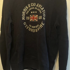 Mörkblå stickad tröja Morris & Co - Snygg mörkblå stickad tröja från Morris & Co med tryck och broderad Union Jack på bröstet. Tröjan har ribbade muddar vid ärmslut och nederkant samt långärmad passform.