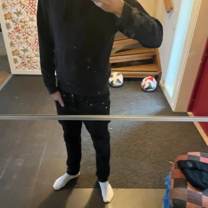 Svart hoodie från Kappa  - Svart hoodie från Kappa i storlek XS med stort gult Kappa-tryck på ryggen. Tröjan har huva med snörning och ribbade muddar vid ärmslut och nederkant. Tillverkad i mjuk bomullsmix, perfekt för chill eller sport. Klassisk streetstil med loose passform.