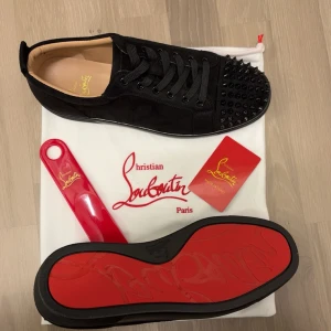 Christian Louboutin svarta sneakers med nitar - Svarta sneakers från Christian Louboutin i mocka med ikonisk röd sula och svarta nitar på tån. Skorna har snörning, rund tå och platt sula. Insidan är i beige skinn och detaljerna är helsvarta för en stilren look.