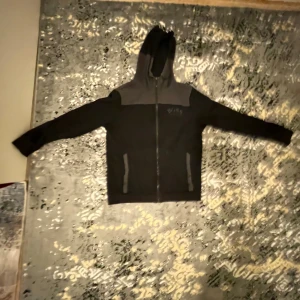 Svart hoodiejacka från BOSS - Svart och mörkgrå hoodiejacka från BOSS med dragkedja framtill och huva med snörning. Jackan har två fickor med dragkedja och en diskret BOSS-logga på bröstet. Perfekt för en avslappnad och stilren look.