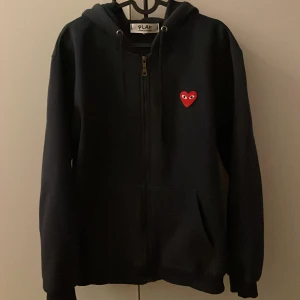 Navy hoodie från Comme des Garçons Play - Navy hoodie med dragkedja och huva från Comme des Garçons Play. På bröstet sitter det ikoniska röda hjärta med ögon. Tröjan har ribbade muddar, fickor fram och är tillverkad i mjuk bomull. Perfekt för dig som gillar streetwear och vill ha något som sticker ut.