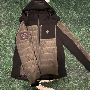 Svart och grön pufferjacka Canada Snow - Snygg pufferjacka från Canada Snow i svart och grönt med quiltad framsida och ärmar. Jackan har huva, dragkedja och coolt lönnlövsmärke på bröstet. Perfekt för kalla dagar och har en modern, sportig look. Flera praktiska fickor och detaljer som gör den extra användbar. Nypris: 3600 Vid mer funderingar eller frågor skriv!