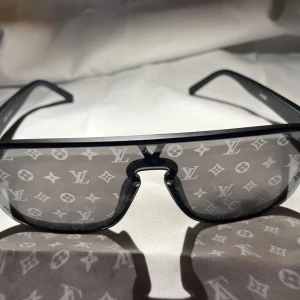 Louis Vuitton svarta monogram-solglasögon - Snygga svarta solglasögon från Louis Vuitton med breda skalmar och spegellins täckt av det ikoniska LV-monogrammet. Sköldpaddsinspirerad båge i modern, sportig form som verkligen sticker ut. Perfekta för dig som vill ha en lyxig och trendig accessoar.