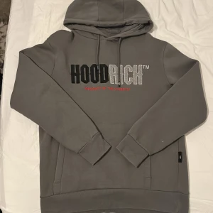 Grå hoodie från Hoodrich XS - Snygg grå hoodie från Hoodrich i storlek XS. Tröjan har broderad logga i svart och silver på bröstet, snörning vid huvan och en klassisk känguruficka framtill. Mjuk insida och ribbade muddar vid ärmar och nederkant.