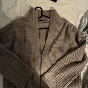 Beige stickad kofta med dragkedja - Mysig beige stickad kofta från lager157 med hög krage och dragkedja framtill. Koftan har en ribbad struktur och långa ärmar, perfekt för lager-på-lager under kyliga dagar.