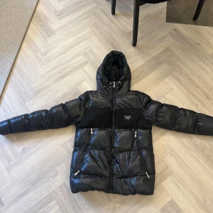 Svart pufferjacka Alessandro Zavetti S - Snygg svart pufferjacka från Alessandro Zavetti med glansig finish, huva och två dragkedjeförsedda fickor. Jackan har en svart panel över bröstet med metallogo och är perfekt för kalla dagar. Stängs med dragkedja framtill.