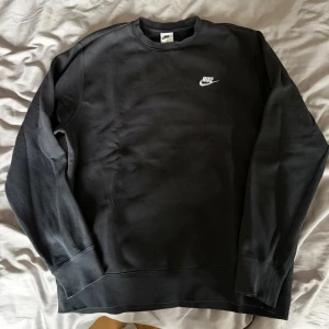Svart sweatshirt från Nike i medium - Svart crewneck sweatshirt från Nike med vit logga på bröstet. Tröjan har rund hals, långa ärmar och ribbade muddar vid ärmslut och nederkant. Tillverkad i mjuk bomullsmix som är skön att ha på sig. Perfekt för chill eller träning.