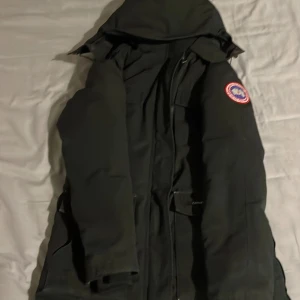 Svart dunparkas från Canada Goose - Säljer en svart parkas från Canada Goose med huva och klassisk logga på ärmen. Jackan är lång, har flera fickor och dragsko i midjan. Perfekt för kalla dagar och riktigt snygg streetstil. Passar dig som vill ha både värme och stil.