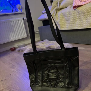 Svart axelväska med flätad detalj - Snygg svart axelväska i skinn med långa remmar och flera praktiska fack med dragkedja. Framsidan har en cool flätad detalj som ger väskan extra edge. Perfekt storlek för att få plats med allt du behöver och enkel att matcha med din outfit.