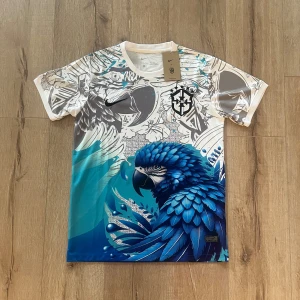 Nike x CBF Dri-Fit t-shirt med papegojtryck - Unik t-shirt från Nike x CBF med ett stort, detaljerat blått papegojtryck och grafiska mönster i grått och vitt. Kortärmad modell i lätt polyester Dri-Fit material som andas. Snyggt CBF-emblem på bröstet och coola kontrasterande färger som sticker ut.