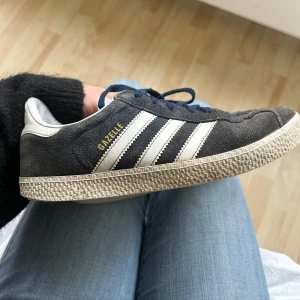 Adidas Gazelle mörkblå sneakers - Säljer ett par klassiska Adidas Gazelle sneakers i mörkblå mocka med vita detaljer och snörning. Skorna har de ikoniska tre ränderna på sidan och en platt, mönstrad sula. Perfekt för dig som gillar retrostil och streetwear.