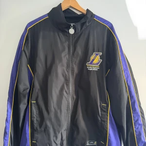 Lakers vindjacka svart/lila gul detalj - Snygg vindjacka från Los Angeles Lakers med broderad logga fram och stort tryck bak. Jackan är svart med lila paneler och gula detaljer längs ärmarna. Dragkedja framtill och hög krage. Perfekt för dig som älskar basket och vill sticka ut.