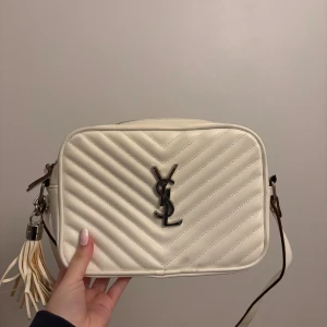 Vit quiltad axelväska från YSL - Snygg vit axelväska från YSL i quiltat skinn med klassisk YSL-logga i metall framtill. Väskan har justerbar axelrem, dragkedja upptill och dekorativ tofs i sidan. Perfekt storlek för det viktigaste och ger en lyxig vibe till din outfit.