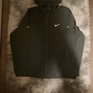 Nike miller  - Svart vindjacka från Nike med huva och hel dragkedja framtill. Jackan har en stilren look med vit Nike-logga på bröstet och diskreta reflexdetaljer på ärmarna. Perfekt för löpning eller utomhusaktiviteter. Tillverkad i lätt polyester som andas.