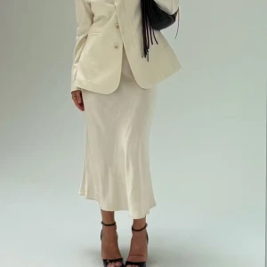 Krämvit satinkjol i lång modell - Supersnygg långkjol i krämvitt satinliknande material med lätt glans. Kjolen har en rak och något utsvängd passform som ger en elegant siluett. Perfekt att styla med kavaj eller stickad tröja för en trendig look.