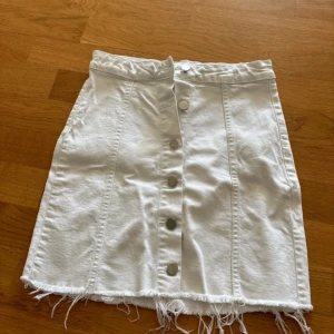 Vit jeanskjol med knappar och fransad kant - Vit jeanskjol med silverfärgade knappar framtill och fransad nederkant. Kjolen är kort och har en rak passform som sitter tajt. 