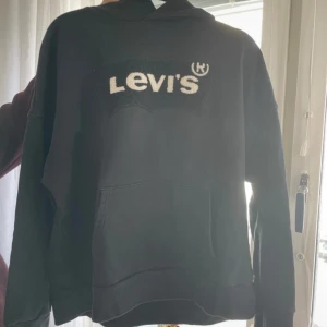 Svart Levi's hoodie med logga - Svart hoodie från Levi's med stor vit logga framtill. Klassisk känguruficka och huva. Tröjan är i mjuk bomull och har långärmad passform, perfekt för chill dagar. Snygg och enkel stil som funkar till jeans eller joggers.