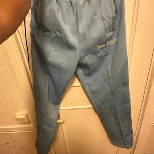 Palm Angels ljusblå mjukisbyxor - Palm Angels ljusblå mjukisbyxor med vita och svarta ränder längs sidorna. Byxorna har elastisk midja, raka ben och logotypen tryckt på framsidan. Perfekt för en chill och sportig look.