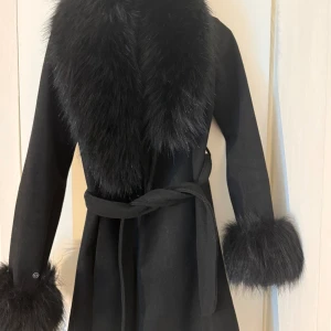 Svart kappa med fuskpälsdetaljer - Stilren svart kappa med bred krage och ärmslut i fluffig fuskpäls. Knytskärp i midjan ger en snygg siluett. Perfekt för dig som vill ha en elegant och trendig look under kallare dagar. Storlek XS Aldrig använd helt ny stått bara i skåpet. 