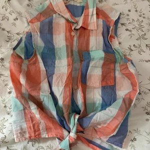 Ärmlös rutig skjorta från H&M - Färgglad ärmlös skjorta från H&M i rutigt mönster med nyanser av blått, korall, mintgrönt och vitt. Skjortan har klassisk krage, knappar framtill, bröstficka och knytning nedtill för en chill vibe. Perfekt för varma dagar och enkel att styla till jeans eller shorts.