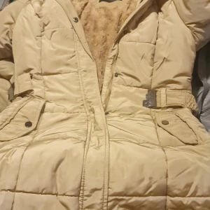 Beige pufferjacka från Glo-Story M - Säljer en beige pufferjacka från Glo-Story i storlek M. Jackan har quiltad design, mjukt pälsfoder på insidan och ett bälte i midjan med metallspänne. Utrustad med praktiska fickor med knappar och dragkedja framtill. Perfekt för kalla dagar.
