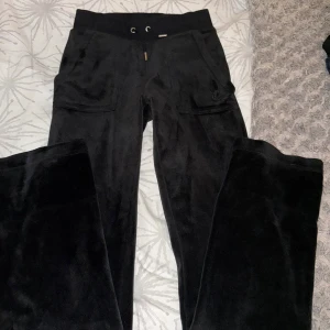 Svarta velourbyxor Juicy Couture XXS - Svarta mjukisbyxor från Juicy Couture i velour med snörning i midjan och fickor både fram och bak. Byxorna har silverfärgade knappar bak och broderad logga på ena fickan fram. Perfekt för en chill och trendig look.