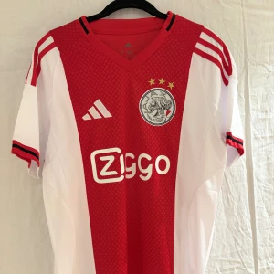 Ajax fotbollströja Adidas S - Ajax hemmatröja från Adidas i slim fit, med klassiskt rött och vitt färgschema, broderat klubbmärke och tre guldstjärnor på bröstet. Tröjan har korta ärmar, V-ringning och Ziggo-logga framtill. Tillverkad i lätt och ventilerande polyester.