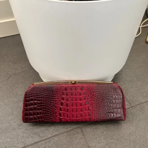 Vintage Röd kuvertväska med krokopräglad yta - Snygg Vintage lite störe röd kuvertväska med krokopräglad yta och guldfärgad metallram. Väskan har ett elegant och avlångt format, med insida i matchande rött tyg den fint skick men är äldre se bilder en fläck. Perfekt för att lyfta din outfit med en vin röd detalj cool detalj och unik struktur. Köpte den på Vinted för 180kr så kan tänka mig sälja den för 150kr?