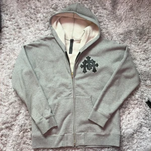 Grå Chrome Hearts hoodie med Silver dragkedja Storlek M - NÄSTAN OANVÄND - Säljer en grå zip hoodie från Chrome Hearts med svart läder i form av kors på bröstet. Tröjan har huva, hel dragkedja med detaljerad silver zip och mjukt, ljust foder. Praktisk känguruficka framtill och ribbade muddar.Väldigt varm! Perfekt för kommande vädret. Perfekt för dig som gillar streetwear med en elegant touch. Storlek M, dock är den på den långre sidan
