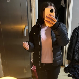 Svart jacka med huva  - Säljer en svart rjacka från vinson med huva och dragkedjor i silver. Jackan har quiltad design, två sidofickor med dragkedja och en extra ficka på ärmen.              