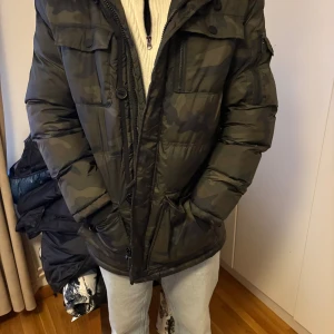 Camouflage pufferjacka med fuskpäls från Ombre - Säljer en mörkgrön camouflage pufferjacka från Ombre med huva och fuskpäls. Jackan har flera fickor med dragkedja och knappar, samt quiltad design. Perfekt för kalla dagar och riktigt snygg streetstil.