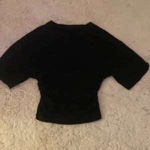 Svart topp med snörning från Gina Tricot - Svart topp från Gina Tricot i storlek S. Modellen har korta ärmar och en snygg snörningsdetalj i sidan som ger en cool och modern look. Materialet är mjukt och stretchigt, perfekt för att skapa en trendig outfit. andvänd några gånger