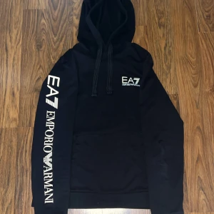  Svart EA7 Emporio Armani hoodie - Svart hoodie från EA7 Emporio Armani med stor vit logga på bröstet och längs ena ärmen. Tröjan har huva med snörning och en klassisk magficka. Mjuk bomullskänsla och långärmad modell, perfekt för en chill och snygg streetstil.