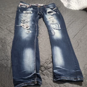 Blå slitna jeans från Jeansnet - Riktigt coola blå jeans från Jeansnet med kraftigt slitna och trasiga detaljer framtill. Jeansen har kontrastsömmar, rutiga detaljer vid fickorna och klassisk femficksdesign. Passformen är rak och materialet är robust denim.