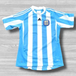 Argentina fotbollströja Adidas herr - Säljer en officiell Argentina landslagströja från Adidas med klassiska ljusblå och vita vertikala ränder. Tröjan har korta ärmar, rund hals och är tillverkad i ventilerande ClimaCool-material. AFA-emblem och Adidas-logga broderade på bröstet. Perfekt för match eller häng med polarna.