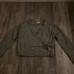 Grå omlottkofta från Kappahl - Mysig grå omlottkofta från Kappahl i storlek 158/164. Koftan har långa ärmar, ribbade muddar och knytband i sidan. Perfekt för lager-på-lager och enkel att matcha med olika outfits.