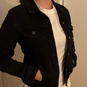 Svart jeansjacka från Bershka M - Svart jeansjacka från Bershka i klassisk modell med knappar framtill och bröstfickor med lock. Snygg passform och perfekt att slänga över en t-shirt. Jackan är i denim och har långa ärmar.