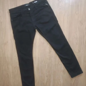 Svarta Replay Anbass jeans slim fit - Svarta jeans från Replay, modell Anbass, med klassisk femficksdesign och diskret Replay-logga på fickan. Jeansen har slim passform och är tillverkade i ett mjukt bomullsmaterial med lite stretch för extra komfort. Perfekta för en clean och stilren look.